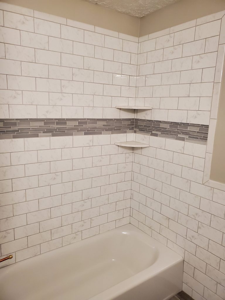 Bathtub surround with marble-look subway tile, glass mosaic accent stripe, and corner shelves